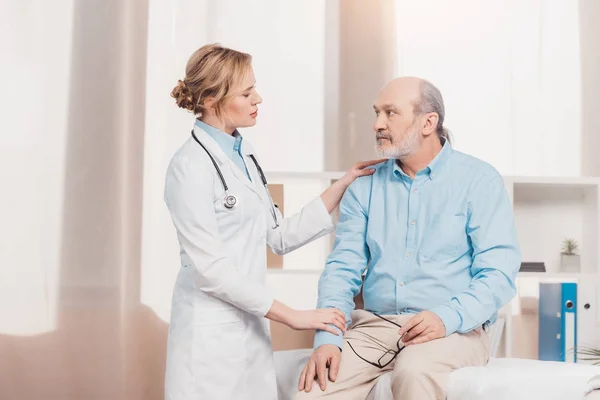 Portrait of doctor in white coat cheering up senior patient in clinic — Stock Photo