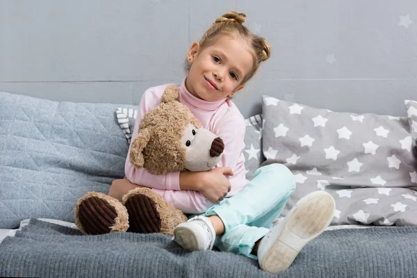 Niño sonriente sentado en la cama con osito de peluche - foto de stock