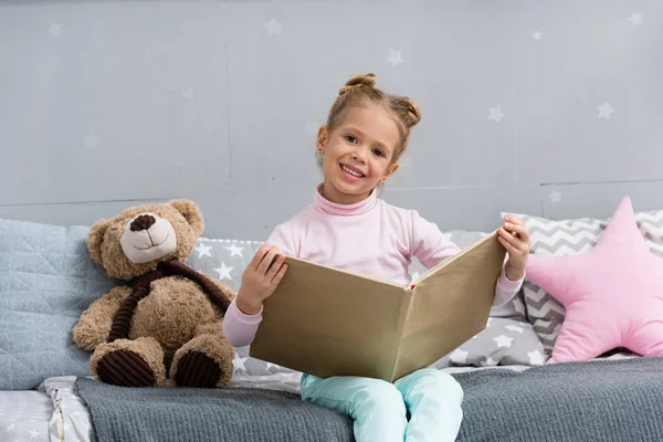 Happy little kid reading book in bed with teddy bear — Stock Photo