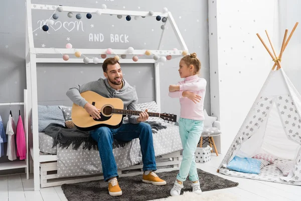 Молодой отец играет на гитаре для дочери в спальне, пока она танцует перед ним — стоковое фото