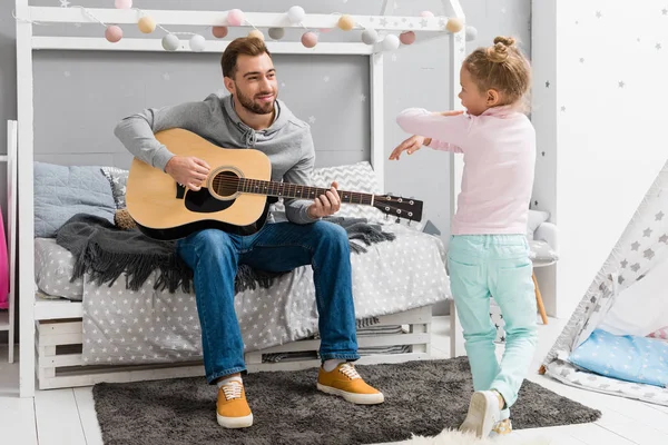 Щасливий батько грає на гітарі для дочки в спальні, поки вона танцює перед ним — стокове фото
