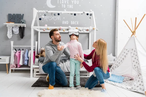 Родители смотрят на маленькую дочь, пока она использует гарнитуру виртуальной реальности в детской спальне — стоковое фото