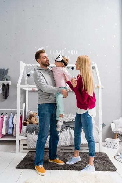 Pais carregando pequena filha enquanto ela usando fone de ouvido realidade virtual no quarto da criança — Fotografia de Stock