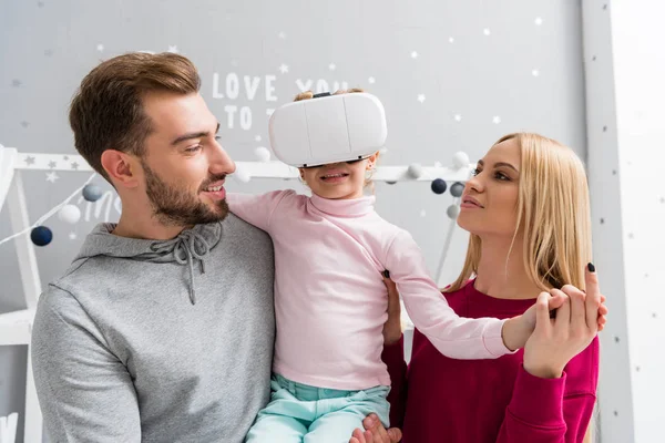 Padres mirando a la pequeña hija mientras ella usa auriculares de realidad virtual - foto de stock