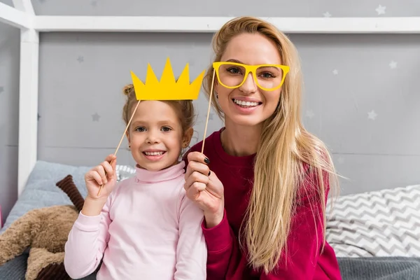 Sonriente madre e hija con juguete mascarada corona y gafas - foto de stock