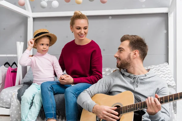 Feliz padre joven tocando la guitarra para la esposa y la hija en el dormitorio del niño - foto de stock