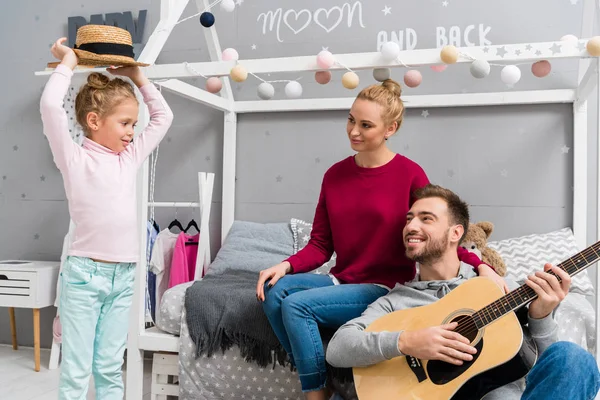 Счастливый молодой отец играет на гитаре для жены и дочери, сидя на полу в детской спальне — стоковое фото
