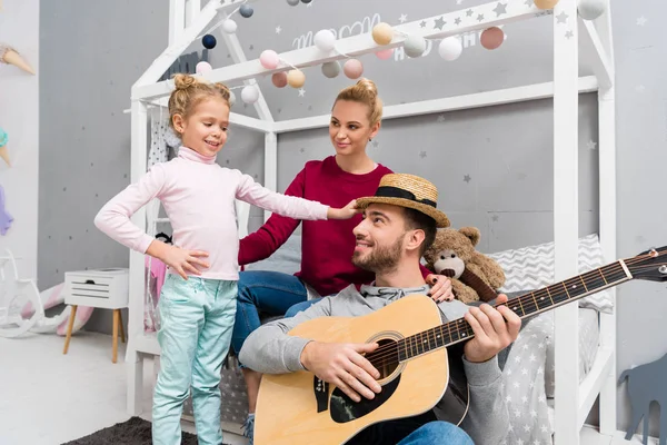 Красивый молодой отец играет на гитаре для жены и дочери в детской спальне — стоковое фото