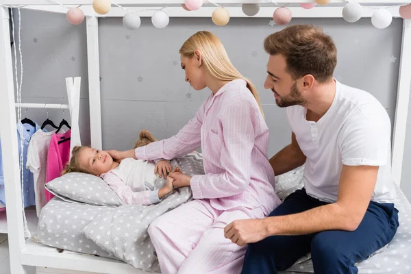 Happy young parents talking to daughter while she lying in bed at kid bedroom — Stock Photo