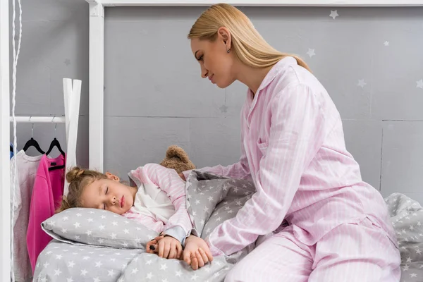 Щаслива мати сидить поруч з дочкою, поки вона спить у ліжку — стокове фото