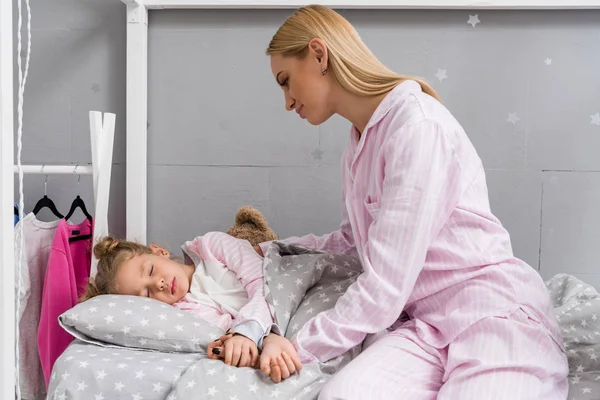 Mère heureuse assise près de sa fille pendant qu'elle dort au lit — Photo de stock