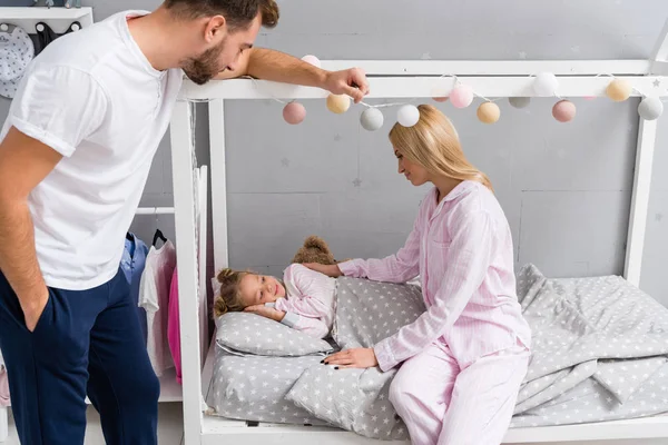 Young parents talking to daughter while she lying in bed at kid bedroom — Stock Photo