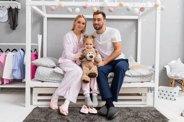 Happy young family sitting on bed in kid bedroom and looking at camera — Stock Photo