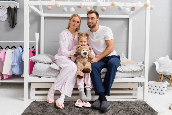 Heureux jeune famille assis sur le lit dans la chambre des enfants et regardant la caméra — Photo de stock