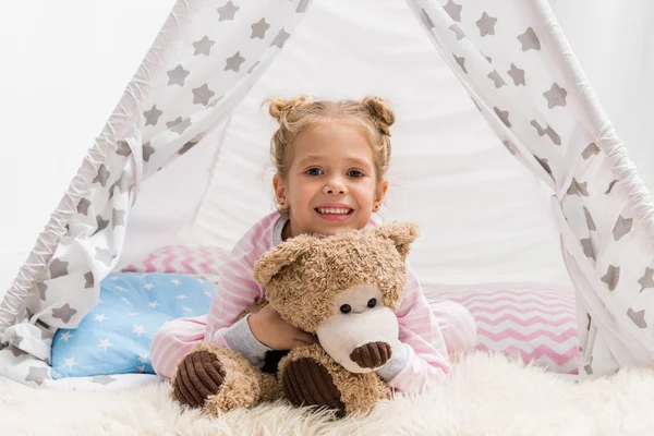 Adorable little kid with teddy bear lying on floor in handcrafted teepee — Stock Photo