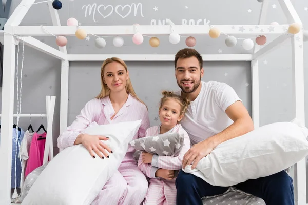 Feliz jovem família sentada na cama com travesseiros e olhando para a câmera no quarto de criança — Fotografia de Stock