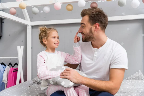 Padre joven y la pequeña hija feliz sentada en la cama en el dormitorio del niño - foto de stock