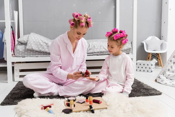 Молода мати малює цвяхи дочки з лаком, сидячи в піжамі з роликами для волосся — стокове фото