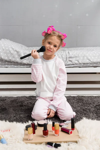 Entzückendes kleines Kind mit Haarrollen auf dem Kopf beim Make-up mit Pinsel, während es auf dem Boden sitzt — Stockfoto