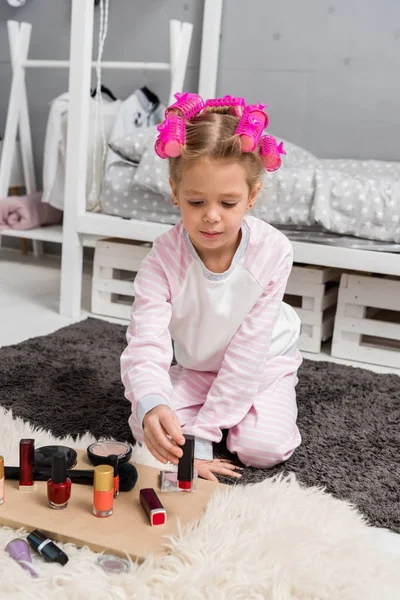 Adorable little kid with hair rollers on head sitting on floor with makeup tools and nail polishes — Stock Photo