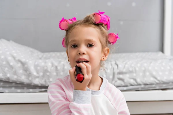 Adorable little kid with hair rollers on head applying lipstick — Stock Photo