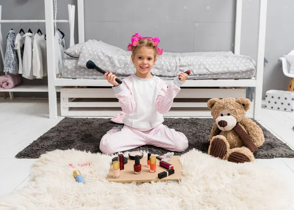 Entzückendes kleines Kind mit Haarrollen auf dem Kopf und Schminkutensilien auf dem Boden sitzend — Stockfoto