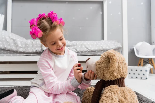 Adorable little kid with hair rollers on head doing makeup for teddy bear — Stock Photo