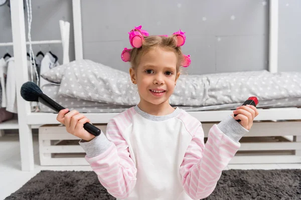 Adorable little kid with hair rollers on head and makeup tools — Stock Photo