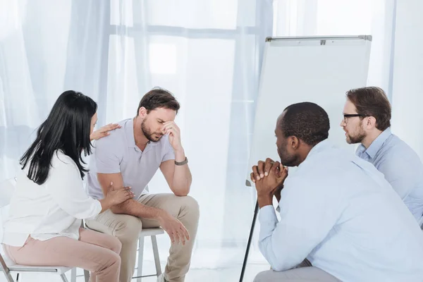 Multiethnic middle aged people sitting on chairs and supporting upset man during anonymous group therapy — Stock Photo