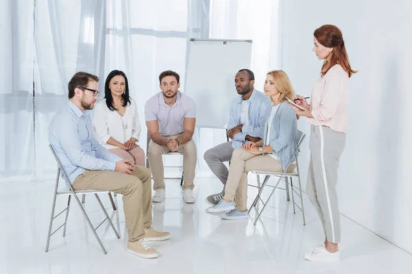 Psicoterapeuta escribiendo en cuaderno y hablando con personas multiétnicas sentadas en sillas durante la terapia de grupo - foto de stock