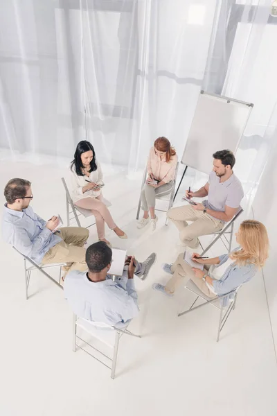 Vista aérea de personas multiétnicas de mediana edad que toman notas en cuadernos durante la terapia de grupo - foto de stock