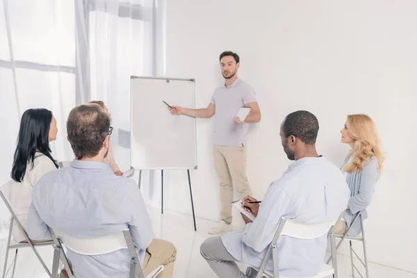 Psicoterapeuta masculino señalando pizarra blanca y grupo multiétnico de personas sentadas en sillas durante la terapia - foto de stock