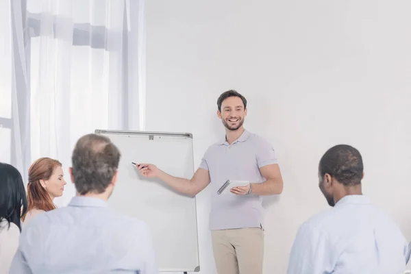 Sonriente hombre sosteniendo el cuaderno y señalando la pizarra blanca en blanco mientras mira a la gente multiétnica durante la terapia de grupo - foto de stock