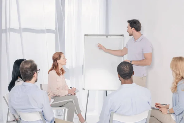 Psychotherapeut zeigt auf leeres Whiteboard und multiethnische Gruppe sitzt während der Therapie auf Stühlen — Stockfoto