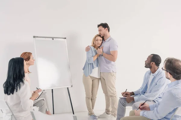 Middle aged couple holding hands while standing near blank whiteboard and other people sitting on chairs during group therapy — Stock Photo