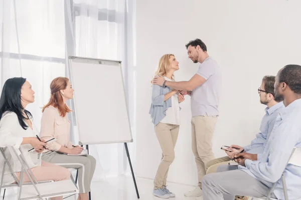 Couple d'âge mûr se regardant tout en se tenant près du tableau blanc blanc et d'autres personnes assises sur des chaises pendant la thérapie de groupe — Photo de stock