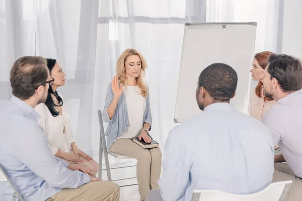 Frau mittleren Alters hält Hand an der Heiligen Bibel, während sie mit multiethnischen Menschen auf Stühlen bei der Gruppentherapie sitzt — Stockfoto