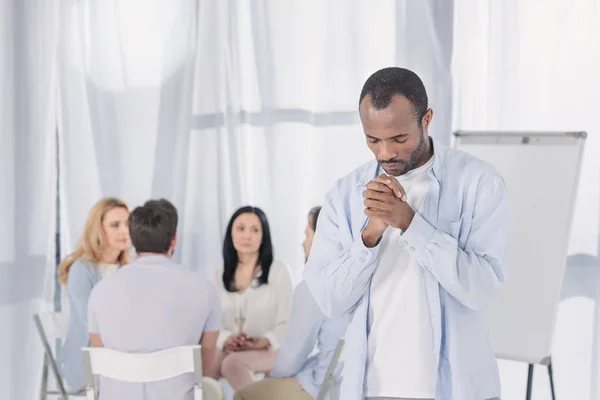 Afroamericano rezando mientras la gente se sienta detrás durante la terapia de grupo - foto de stock