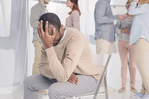 Upset african american man sitting on chair while people standing behind during group therapy — Stock Photo