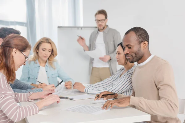 Multiethnic team working with papers together while man standing near whiteboard behind — Stock Photo