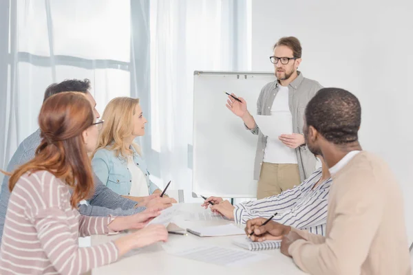 Middle aged man in in eyeglasses with papers standing at whiteboard and having conversation with his team — Stock Photo