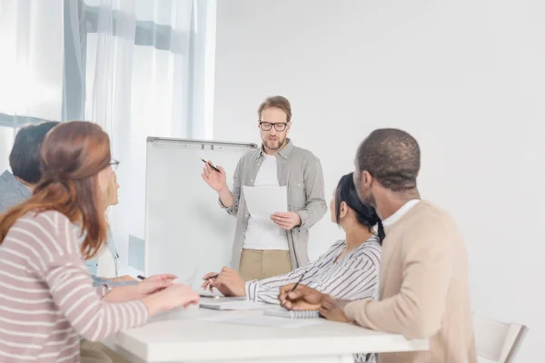 Uomo di mezza età in occhiali in possesso di carta e avendo conversazione con la sua squadra mentre in piedi alla lavagna bianca — Stock Photo