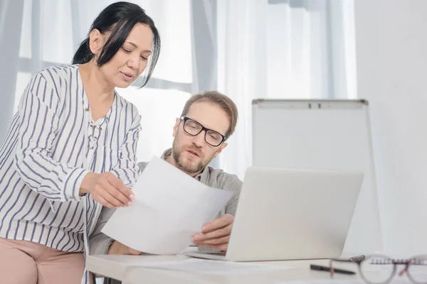 Smiling middle aged businesswoman showing documents to man using laptop in office — Stock Photo