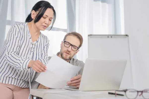 Sonriente mujer de negocios de mediana edad mostrando documentos al hombre usando el ordenador portátil en la oficina - foto de stock