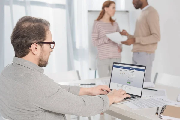 Middle aged man in in eyeglasses using laptop with facebook website on screen in office — Stock Photo