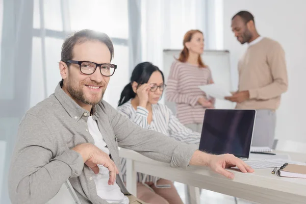 Handsome mature man in eyeglasses smiling at camera while multiethnic people with laptop and papers working behind — Stock Photo
