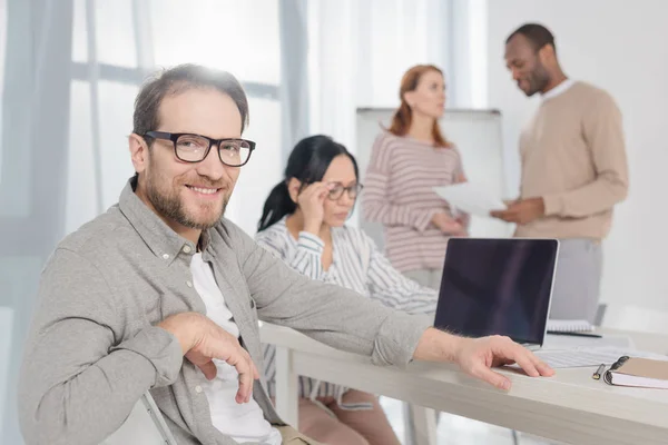 Bell'uomo maturo con gli occhiali che sorride alla macchina fotografica mentre persone multietniche con laptop e carte che lavorano dietro — Foto stock