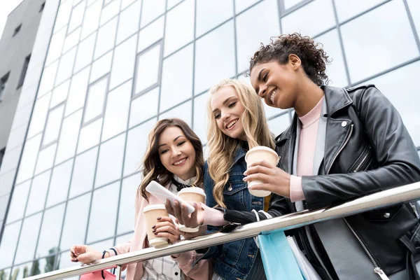 Group of young girlfriends with coffee to go using smartphone together after shopping — Stock Photo