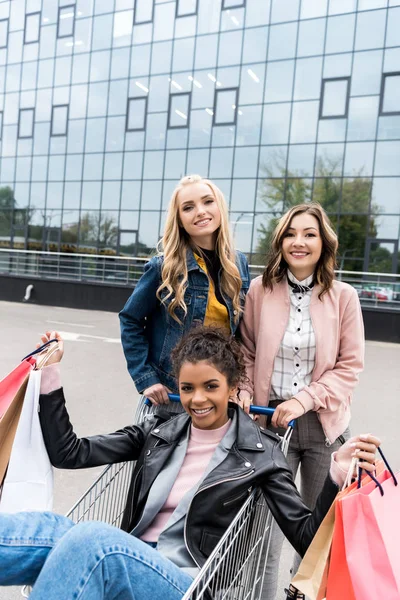 Group of happy young women with shopping cart riding on parking — Stock Photo