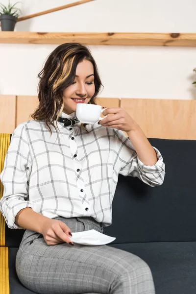Smiling young woman drinking coffee while sitting on couch in cafe — Stock Photo